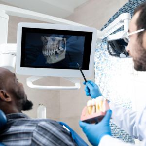 Dentist wearing loupe glasses examines a patients teeth while viewing a dental X ray on a monitor in the dental clinic