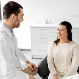 Male doctor in a white coat speaking with a smiling female patient in a bright clinic exam room