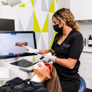Dentist in black scrubs and a mask demonstrates a dental model on a computer screen to a patient lying back in a dental chair