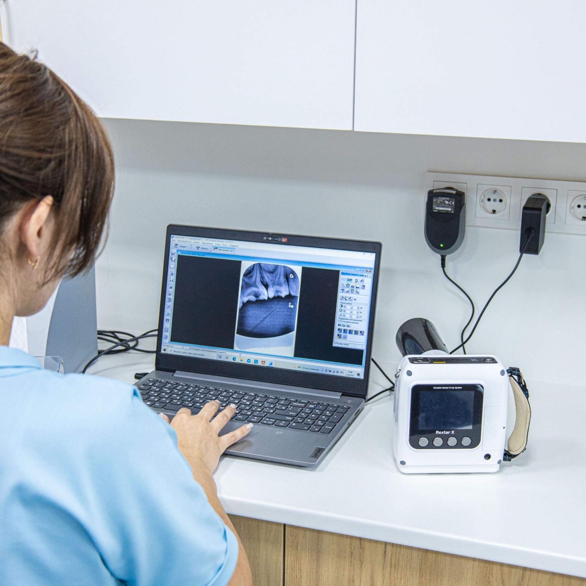 Dental professional in blue scrubs typing on a laptop at a clinic counter with dental equipment nearby