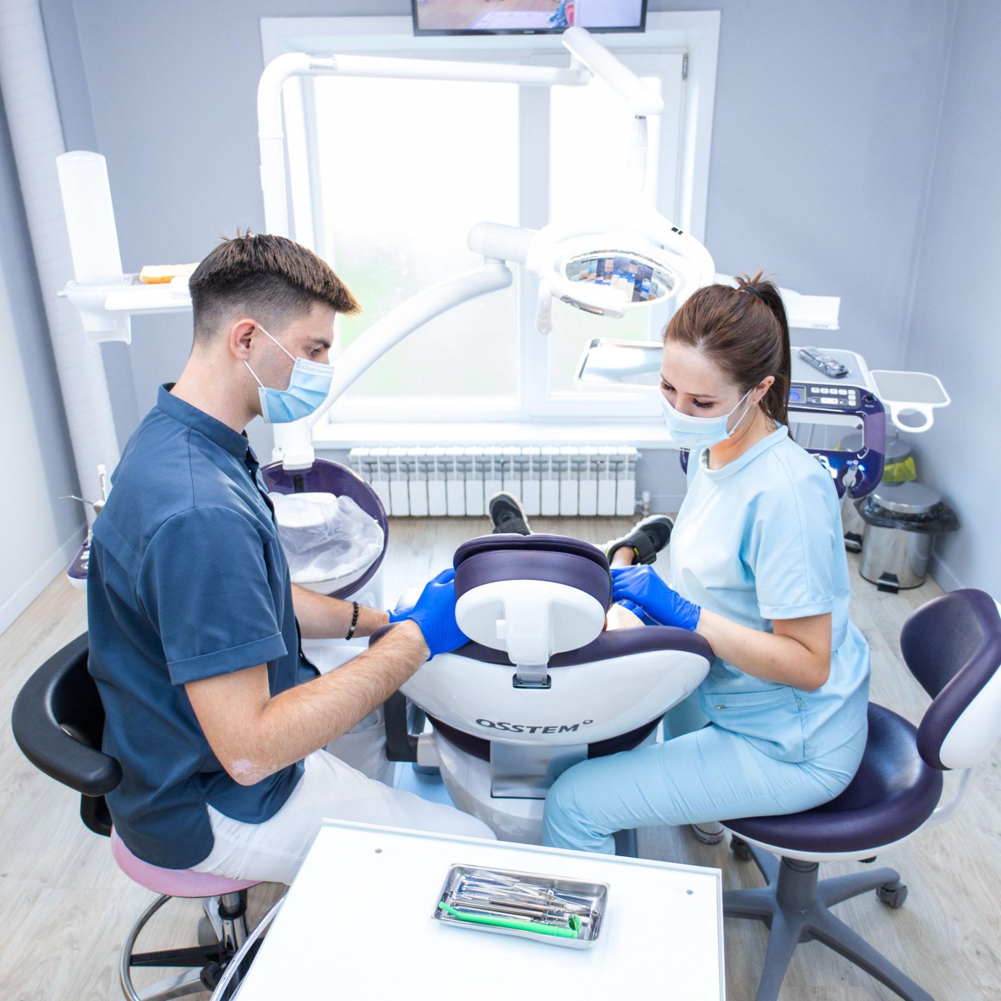 Dentist and dental assistant wearing masks work at a patient chair in a bright dental clinic with instruments nearby
