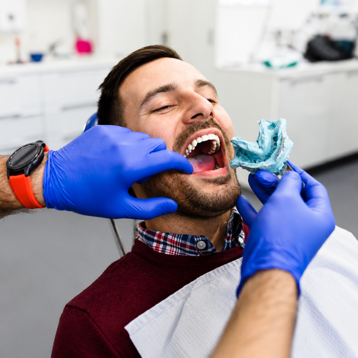 Man in a dental chair with his mouth open while a clinician in blue gloves holds a dental impression tray near his teeth in a bright clinic